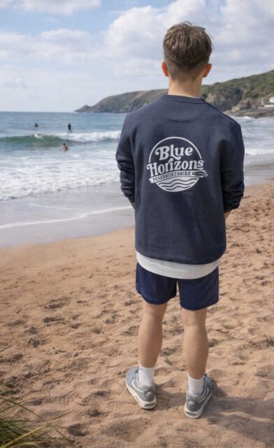 Children from the Blue Horizons kids club run and jump along the coastal dunes overlooking the sea in Pembrokeshire. Wearing Blue Horizons hoodies, they are enjoying time outdoors as part of the inclusive surf community. Blue Horizons provides adaptive surfing, surf therapy and ocean activities designed to help children of all abilities build confidence, friendships and a connection with the sea.