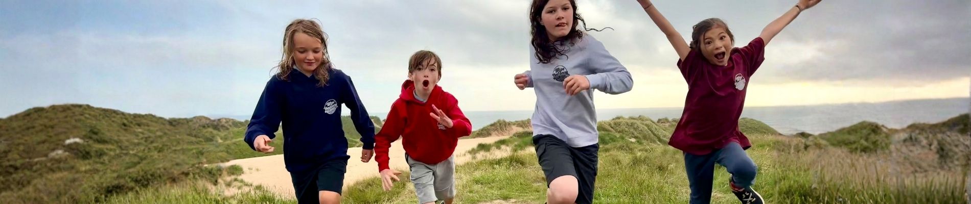 Children from Blue Horizons surf club wearing Blue Horizons hoodies running and jumping on coastal dunes in Pembrokeshire.